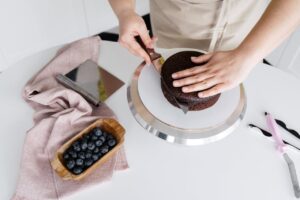 Crop anonymous person cutting delicious homemade cake placed on plate on white table with blueberries and cloth in light kitchen